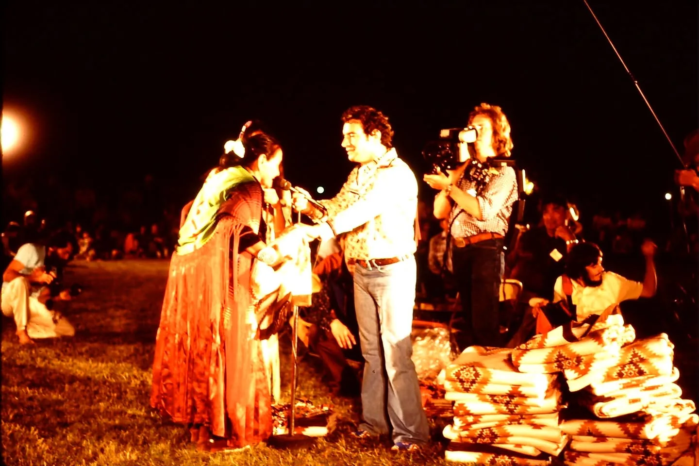 A man with a microphone greets a woman in a fringed shawl beside stacked blankets as a videographer films.