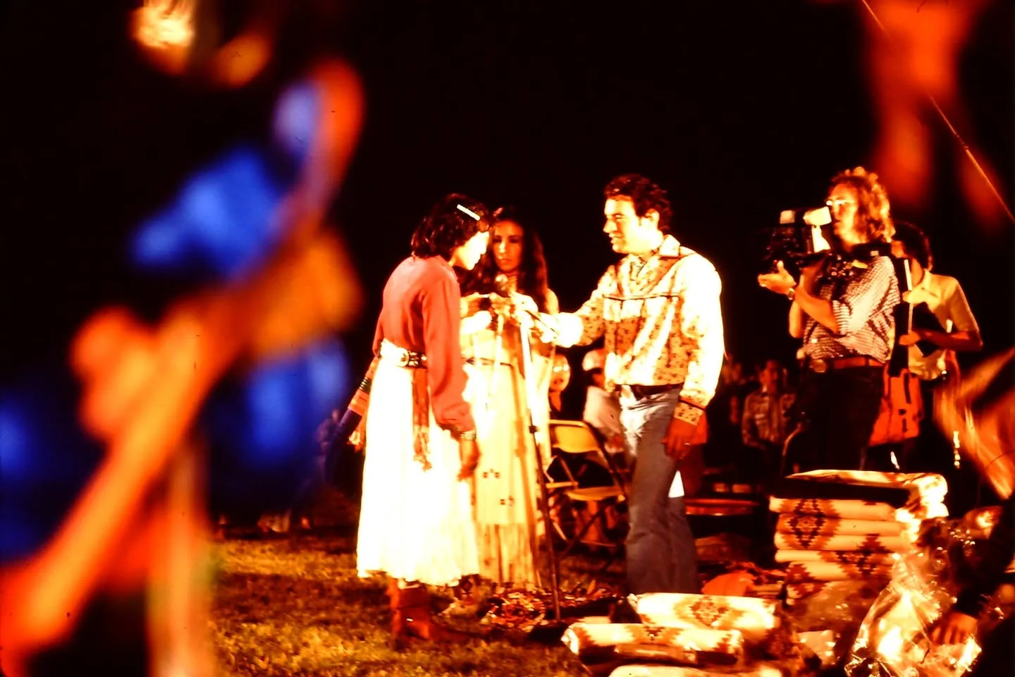 A man holding a microphone stands before two women, one in a white skirt, near stacked blankets at night.