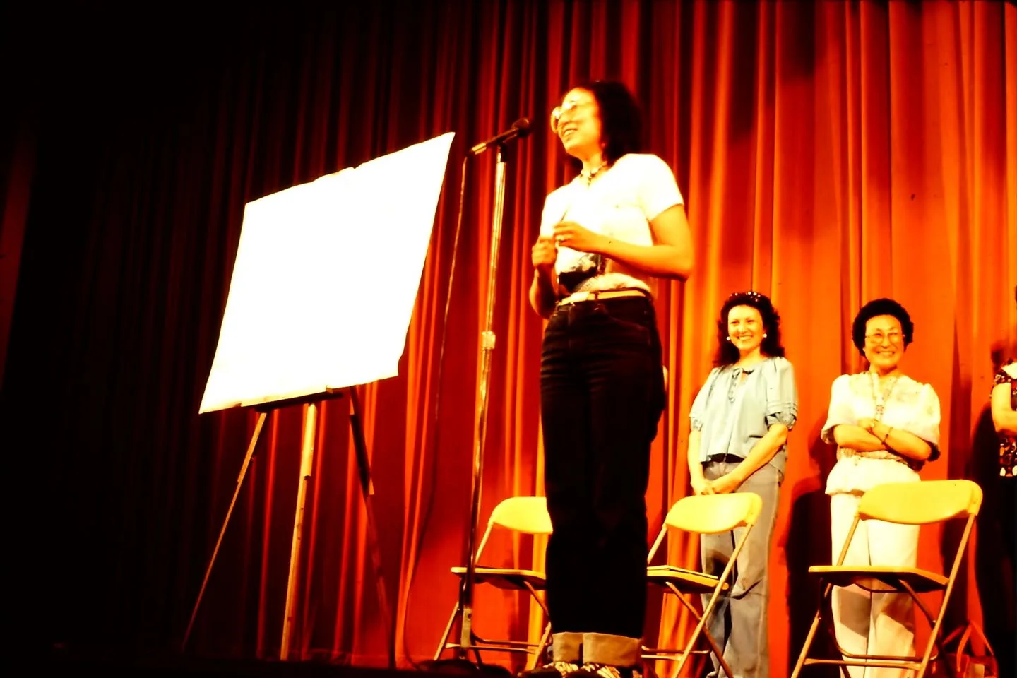 A smiling woman speaks at a microphone on stage beside an easel while two women stand smiling behind her.