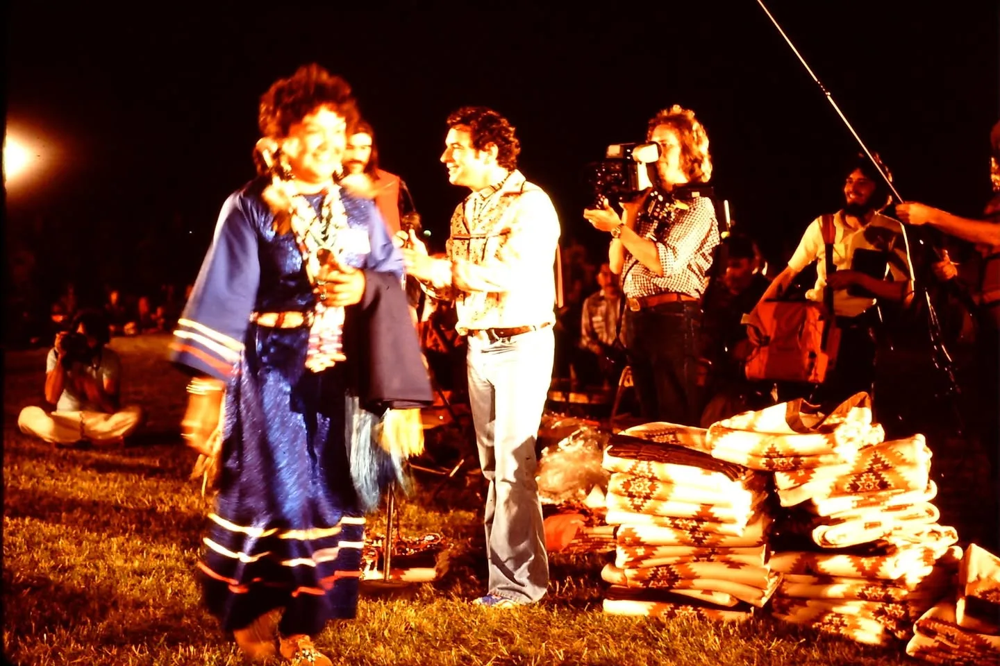 A woman in a blue ribbon dress walks past a man with a microphone and stacks of patterned blankets as a camera operator films.