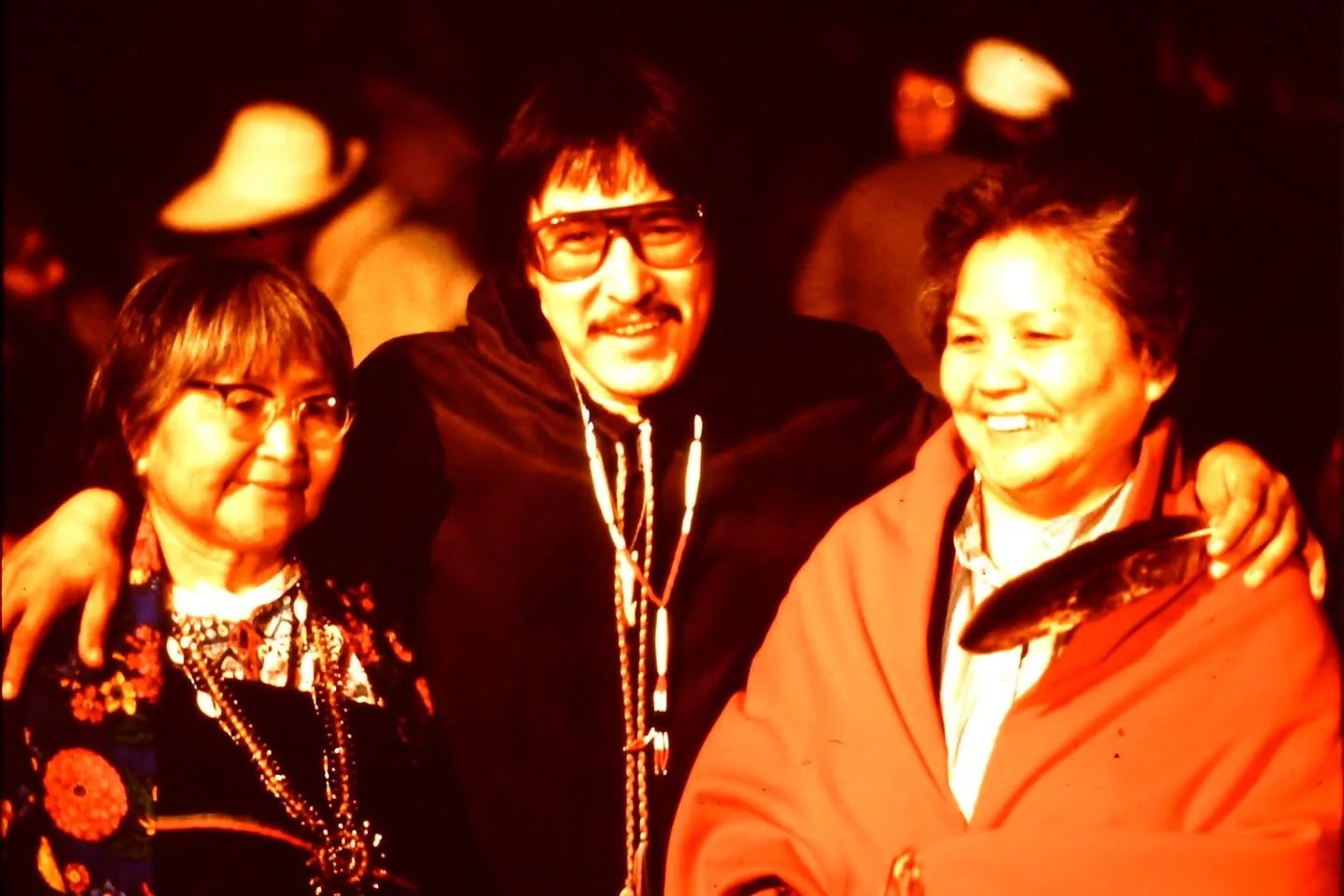 A man in glasses and beaded necklaces puts his arms around two smiling older women at night.