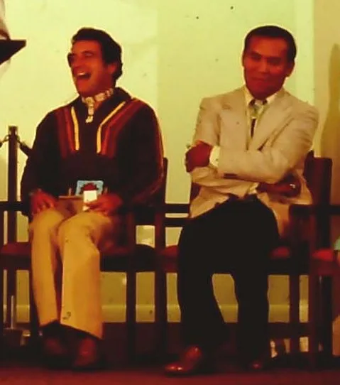 Two men sit side by side, one laughing in a striped shirt, the other in a light suit with arms crossed.