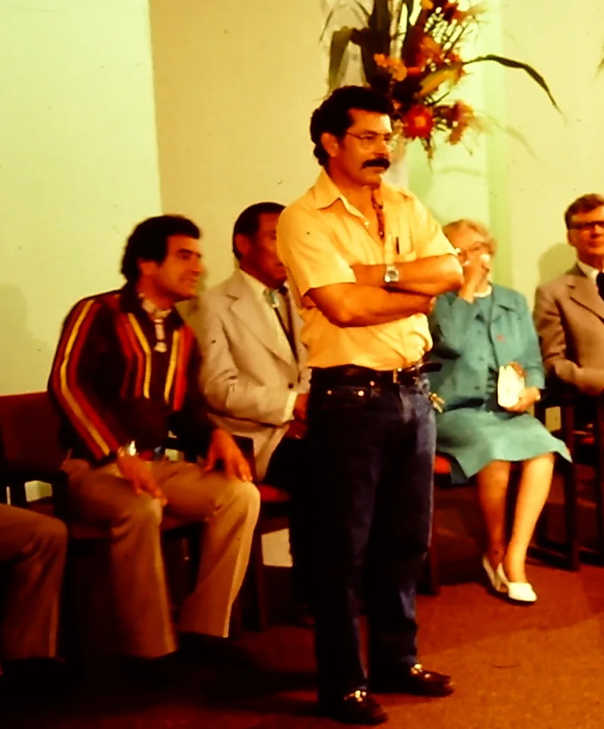 A man in a yellow shirt stands with arms crossed in front of a row of seated people.