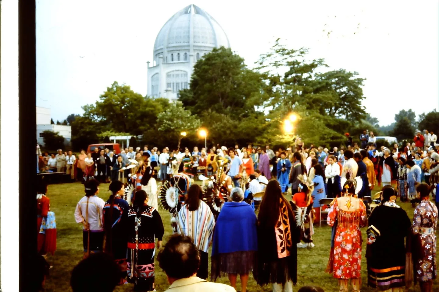 People in regalia and shawls stand in a wide circle on a lawn at dusk, with a white domed temple beyond the trees.