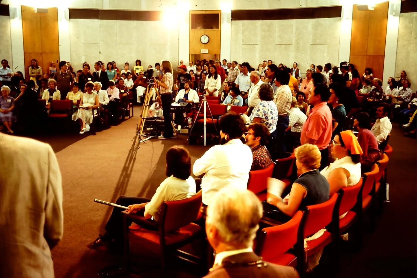 A large audience fills a round hall with red chairs as video cameras on tripods record.