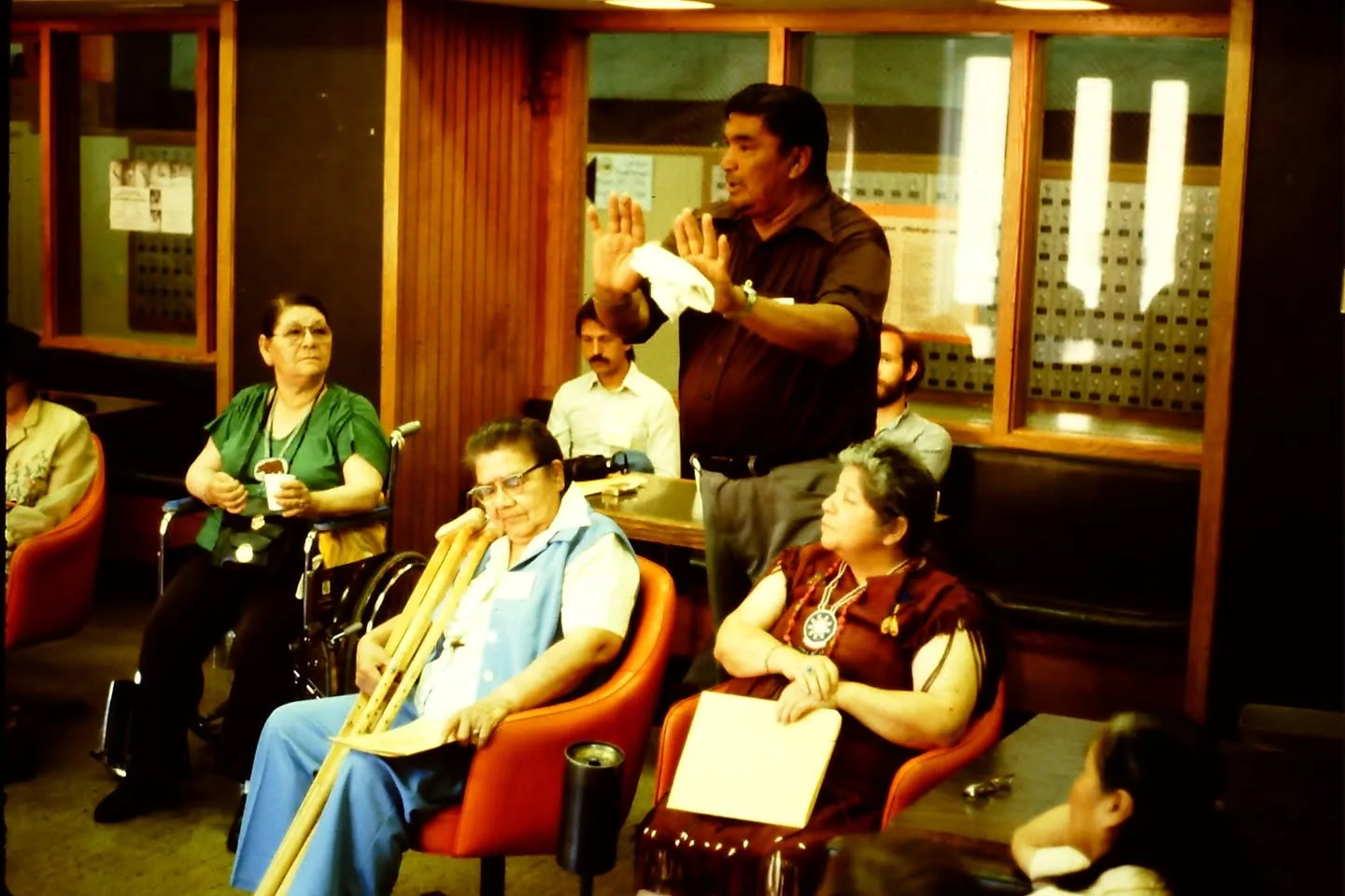A man stands speaking with his hands raised while elders sit listening, one holding crutches and one in a wheelchair.