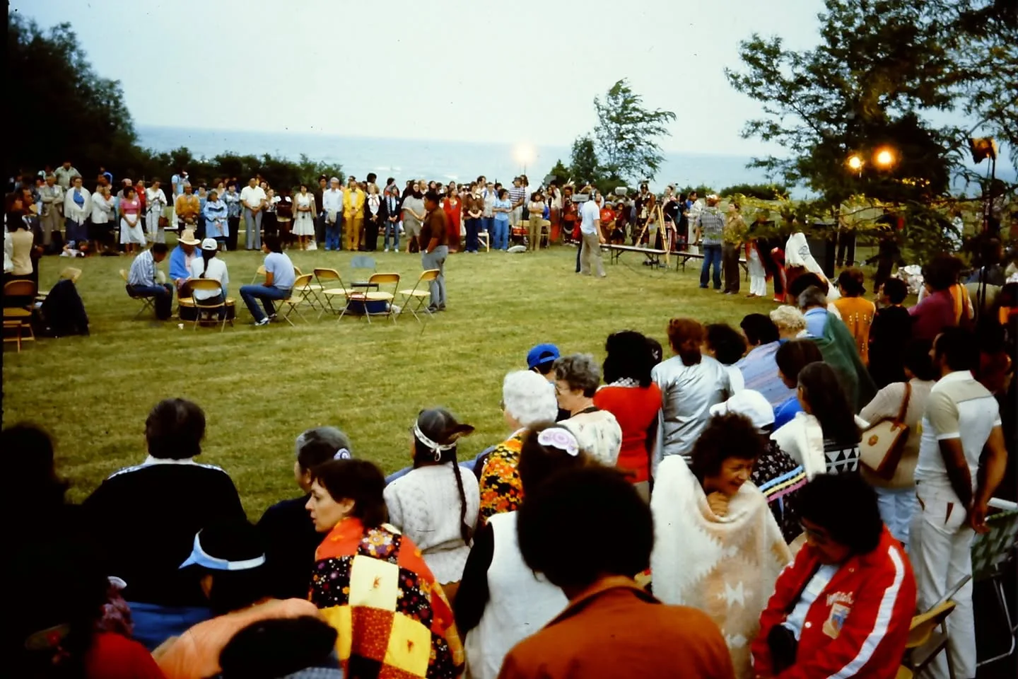 A large circle of people gathers on a lawn beside a lake, with a drum group seated on chairs in the centre.