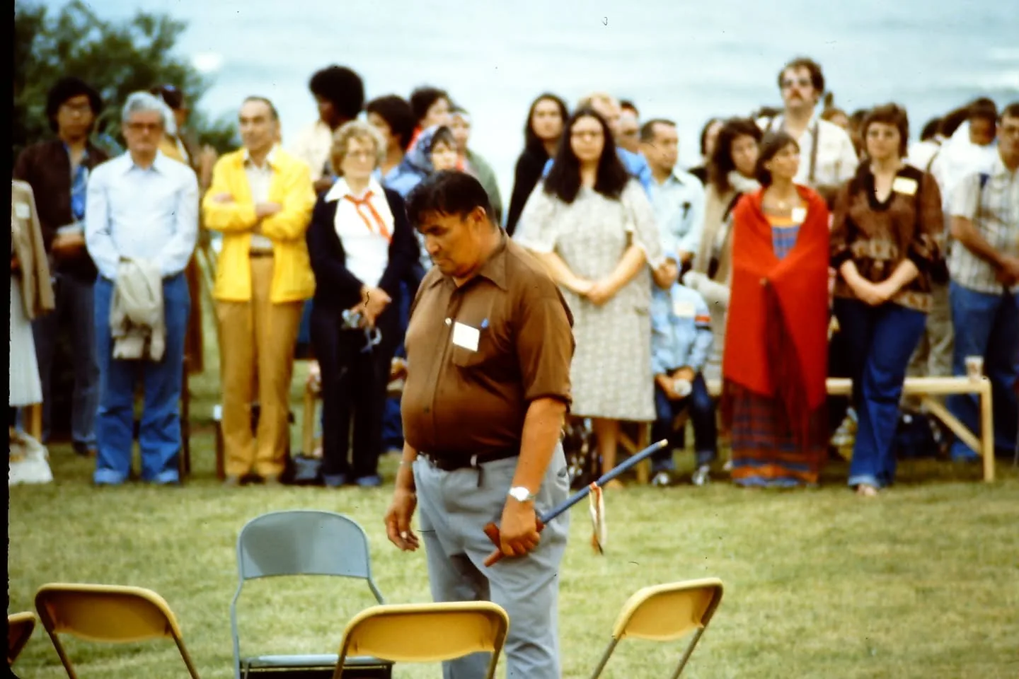 A man in a brown shirt with a name tag carries a decorated staff past folding chairs as a crowd stands watching.