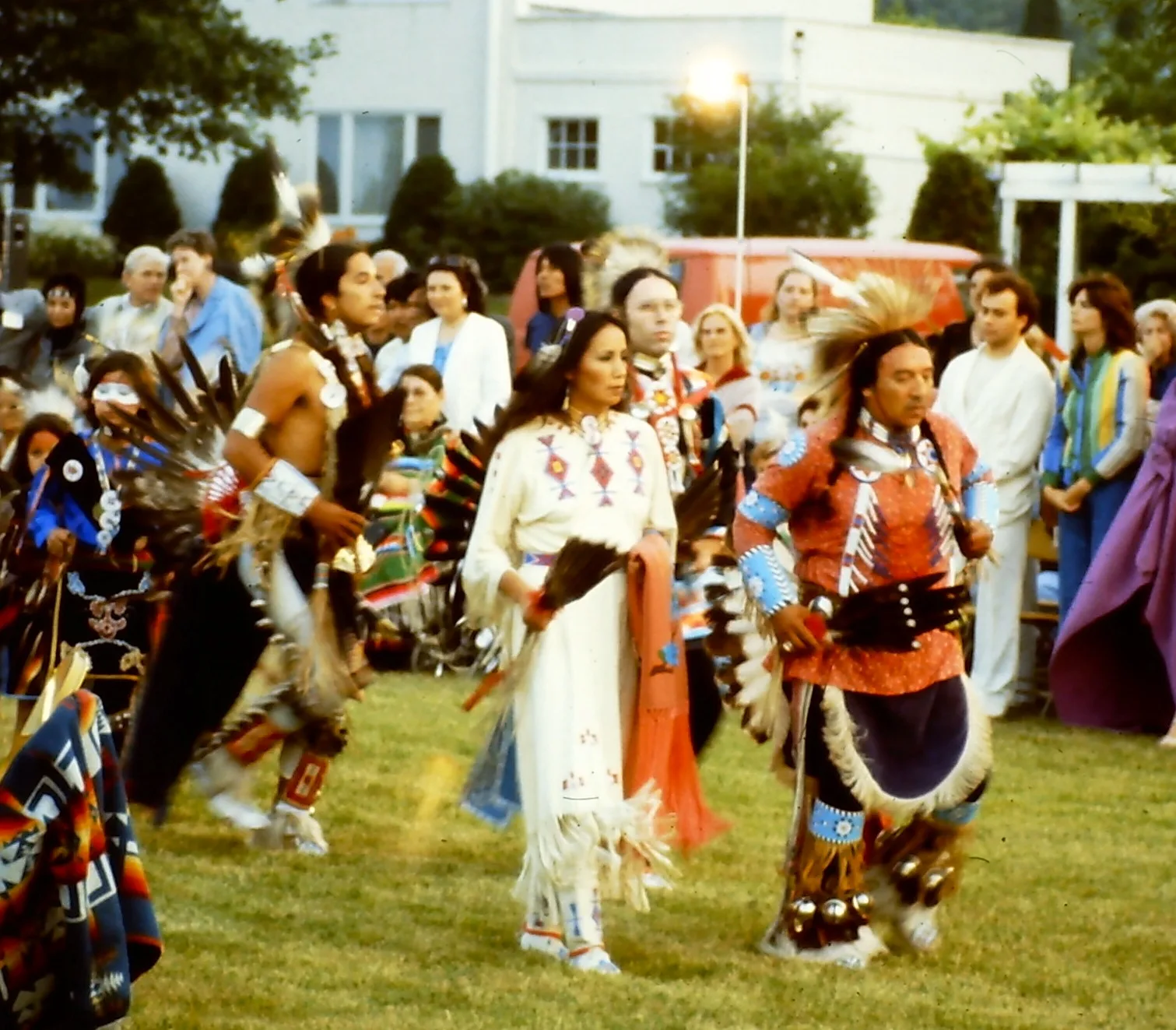 Dancers in regalia move together: a man with feathers, a woman in a white beaded buckskin dress, and a man in a red shirt with a feather bustle.