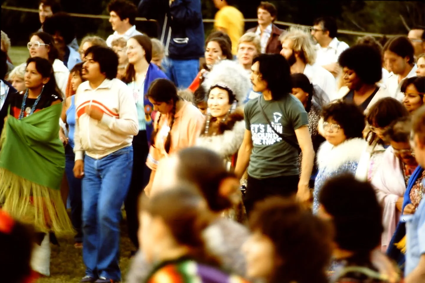 A crowd of people, some in regalia and one woman in a fur-trimmed parka hood, dances together on a field.
