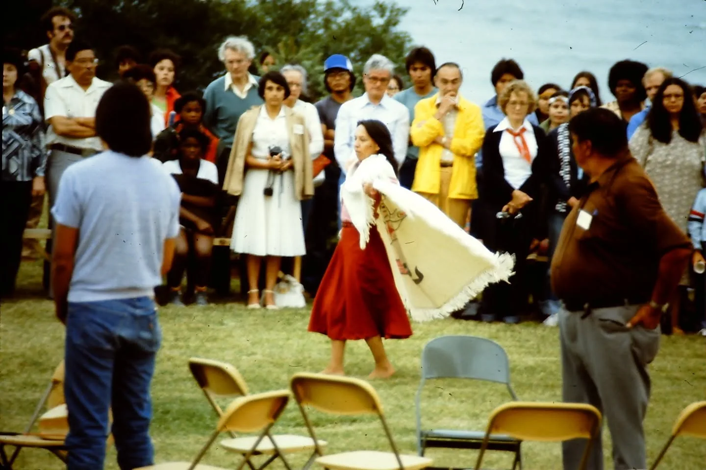 A woman in a red skirt dances barefoot with a white fringed shawl as a crowd stands watching, a lake behind them.