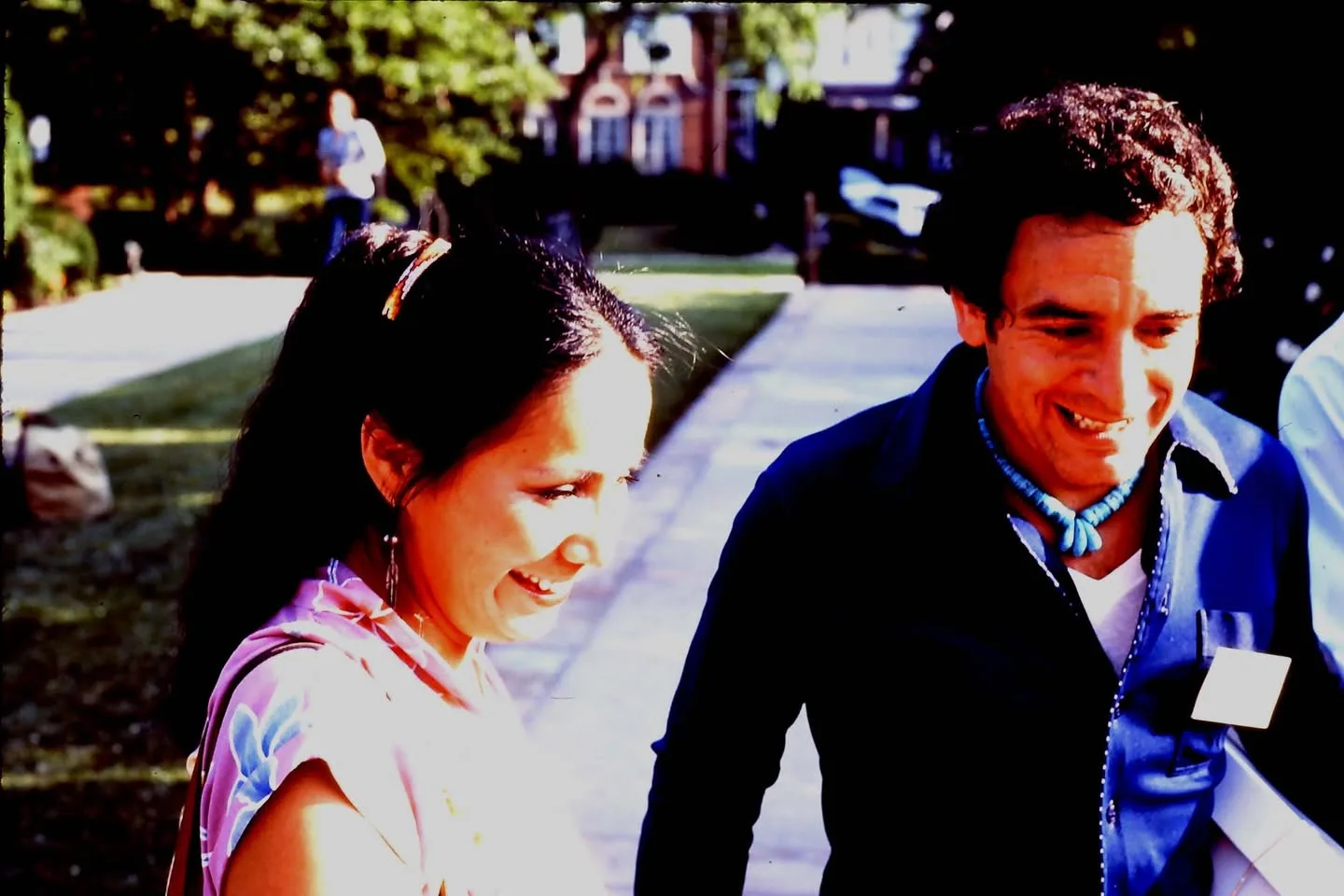 A smiling woman and a smiling man wearing a turquoise necklace stand together on a sunny walkway.