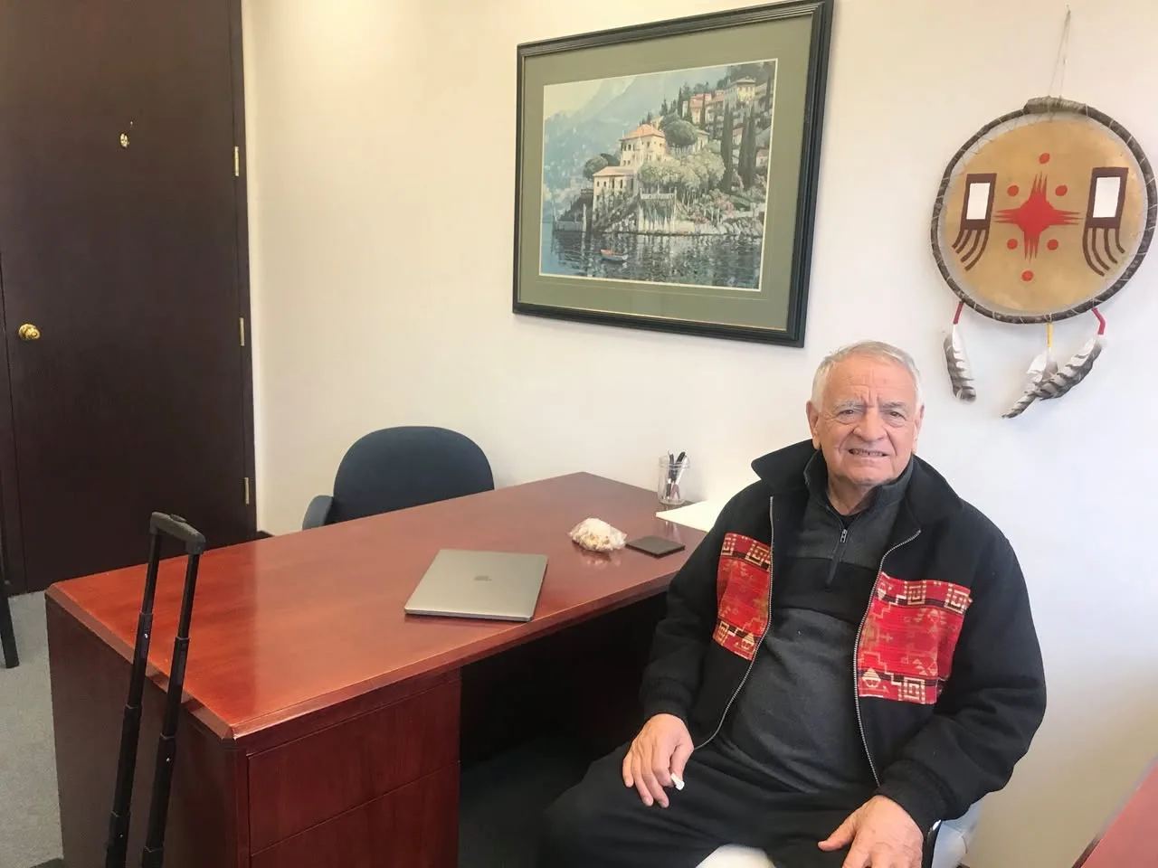 Phil Lane Jr. smiles while seated beside a wooden office desk with a laptop, wearing a black jacket with red patterned panels; a painted hide shield with feathers hangs on the wall.