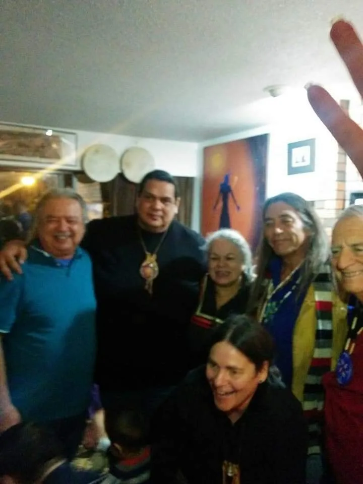 A smiling group of Indigenous elders and relatives pose close together in a home, with hand drums and a painting on the wall behind them.