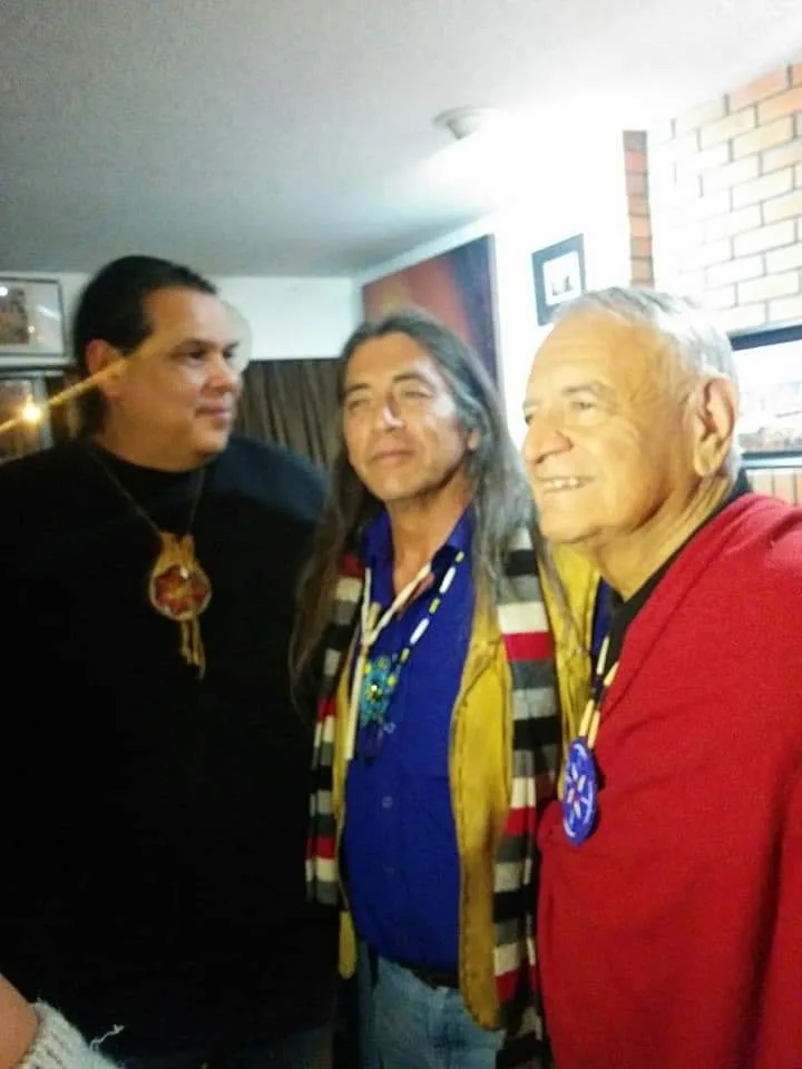 Three men stand together: one in black with a beaded pendant, a long-haired man in a yellow vest and striped sash, and a smiling elder in red.