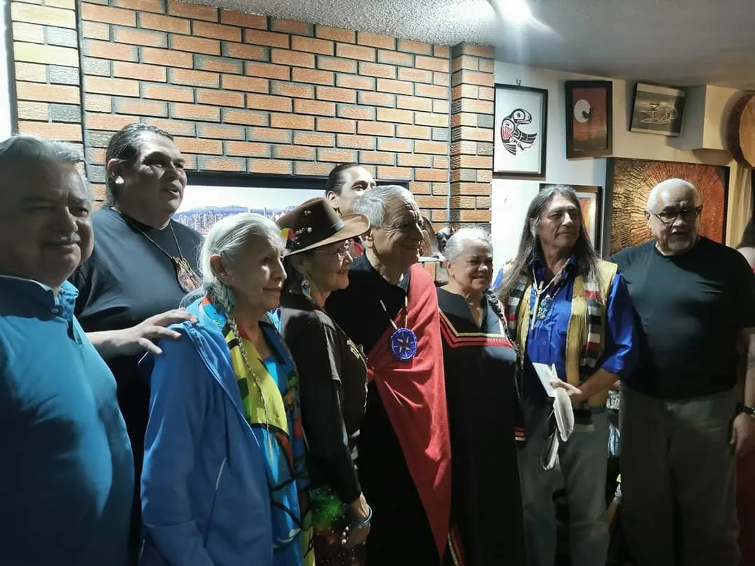 Elders and relatives stand in a row before a brick wall hung with Coast Salish art; an elder at center wears a red blanket and beaded medallion.