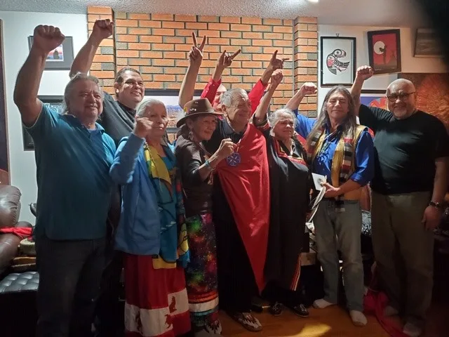 The group raises fists and peace signs in unity around an elder wrapped in a red blanket, in a living room with framed Indigenous art.