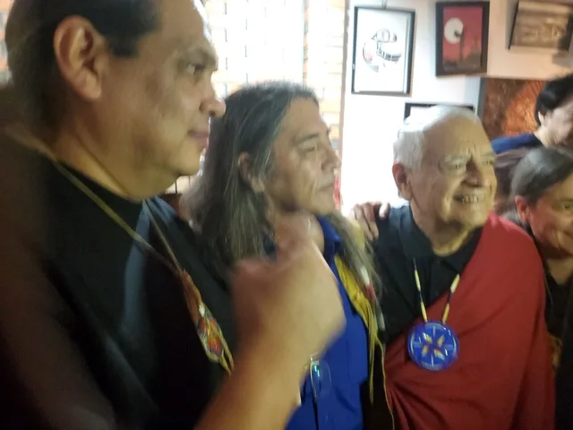 Close-up of three men side by side; a white-haired elder in a red blanket and blue beaded medallion smiles.