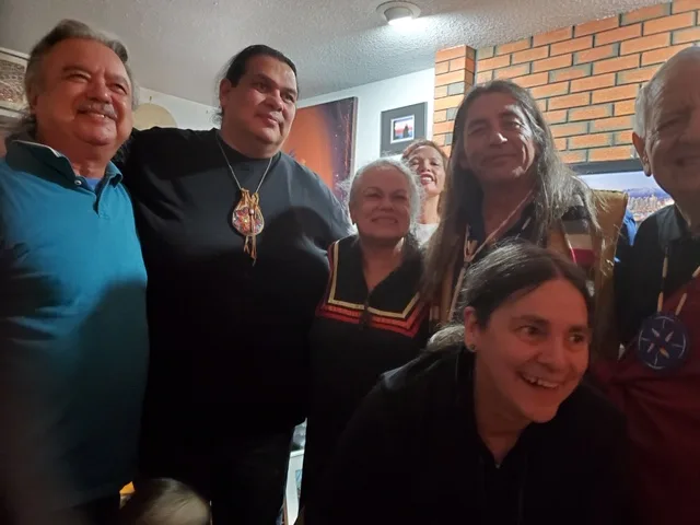 A smiling group of elders and relatives crowd together in a home, with a woman crouched in front and a brick wall behind.