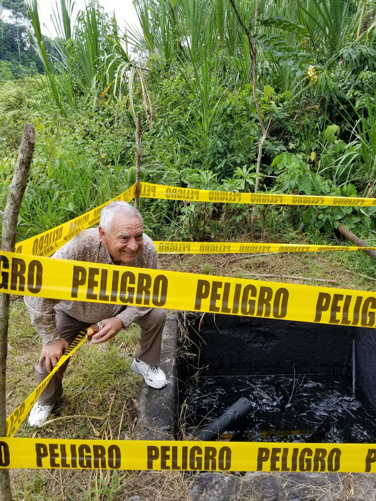 A smiling white-haired elder in a patterned shirt crouches beside a pit of black oil sludge cordoned off with yellow PELIGRO tape in the jungle.