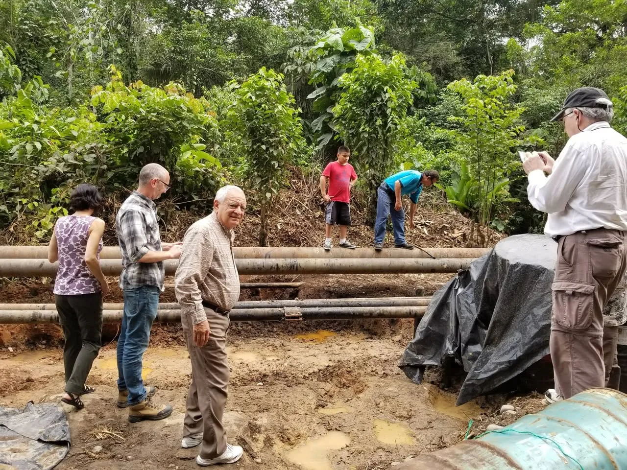 A small group, including the white-haired elder, stands on muddy ground near large oil pipelines in the forest while others inspect the site and take photos.