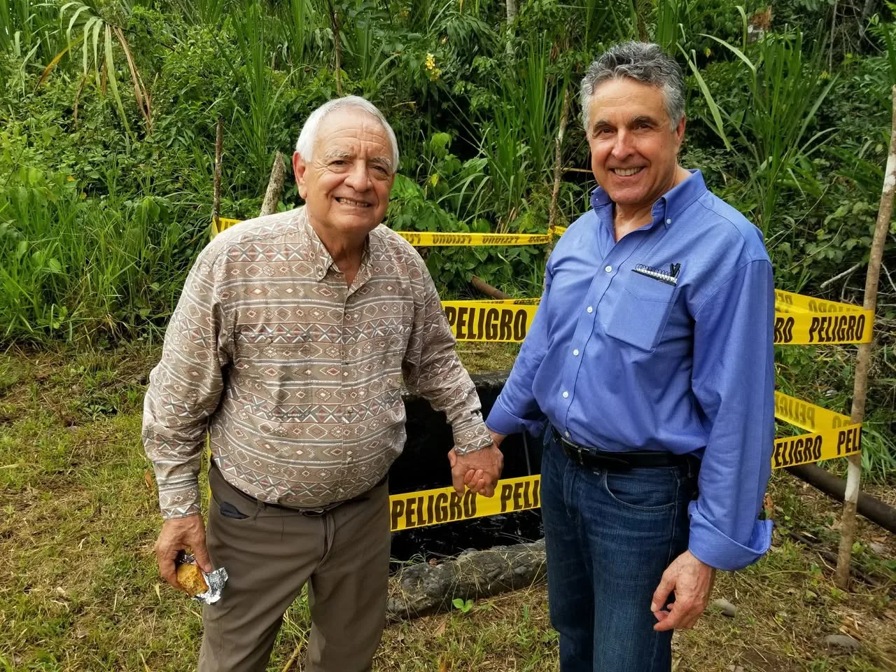 Phil Lane Jr. and Charles McNeill hold hands and smile in front of an oil pit surrounded by yellow PELIGRO danger tape.