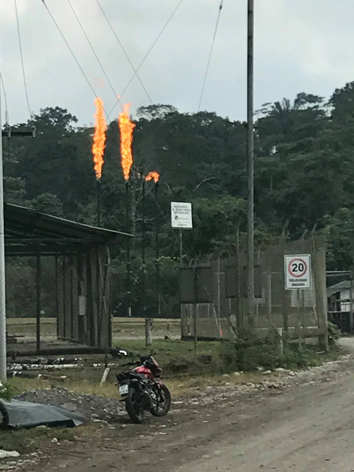Tall flames burn from gas flaring pipes against a forested hillside, behind a fence near a dirt road and a parked motorcycle.