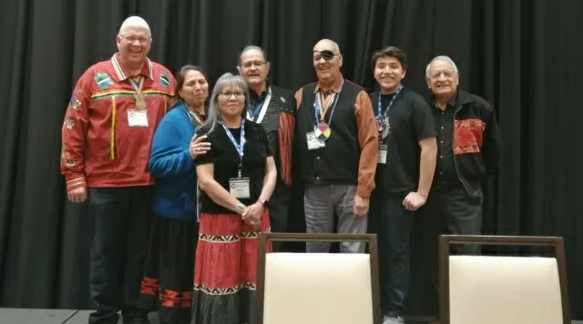 Seven people stand together on a stage before a black curtain, wearing conference lanyards; several elders wear ribbon shirts and a young man stands among them.