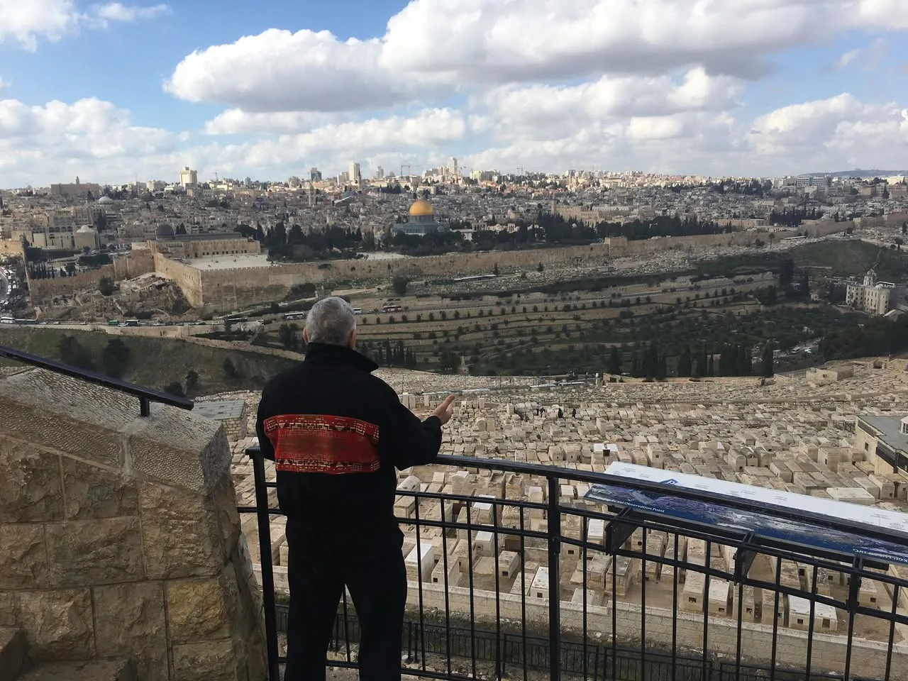 An elder with grey hair in a black jacket with a red patterned band stands at a railing, gesturing toward Jerusalem's Old City, the Dome of the Rock and a vast cemetery below.