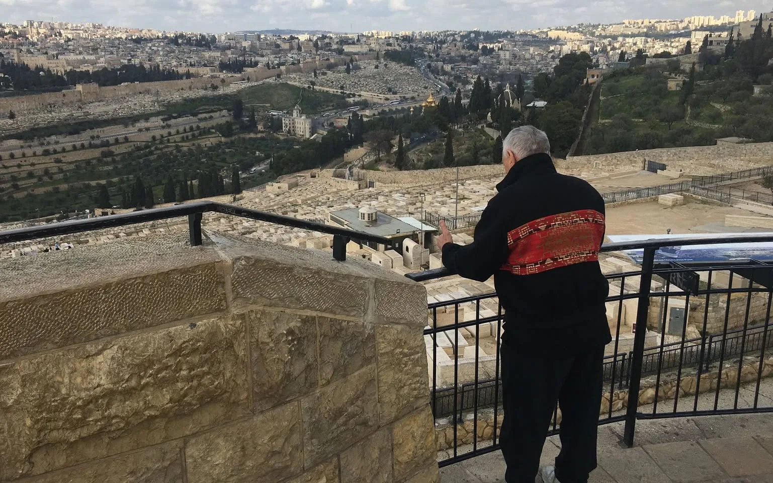 The same elder leans on a railing beside a stone wall, looking out over the terraced Jewish cemetery on the Mount of Olives and the city beyond.