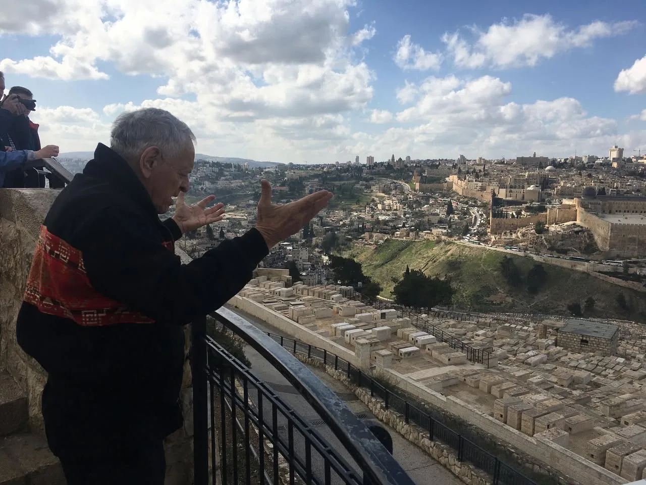 The elder raises both open hands in prayer at the railing, overlooking rows of stone tombs and the walls of Jerusalem under a cloudy blue sky.