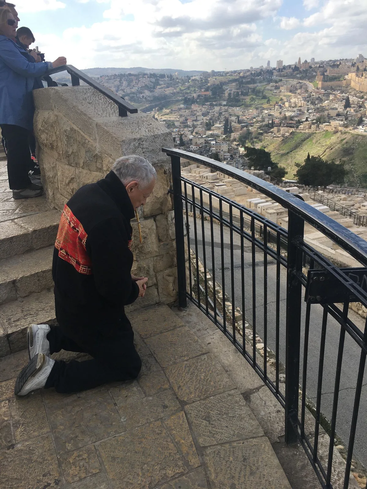 The elder kneels in prayer on a stone overlook with hands clasped, holding a small object to his lips, as visitors stand nearby above the cemetery.