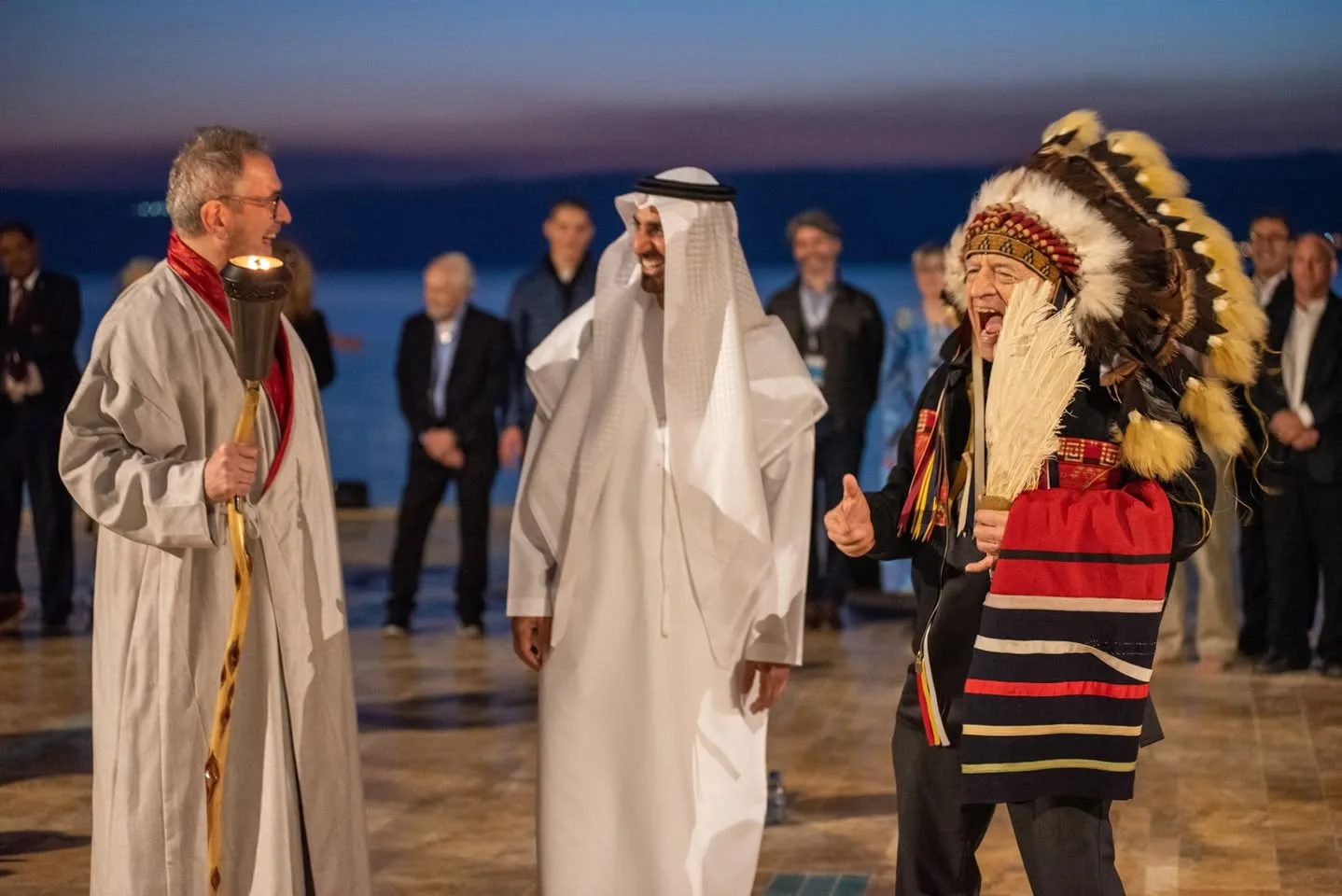 The elder in the feathered headdress laughs and gives a thumbs-up while the man in white robes and the torch bearer share the laughter, with a circle of guests behind.