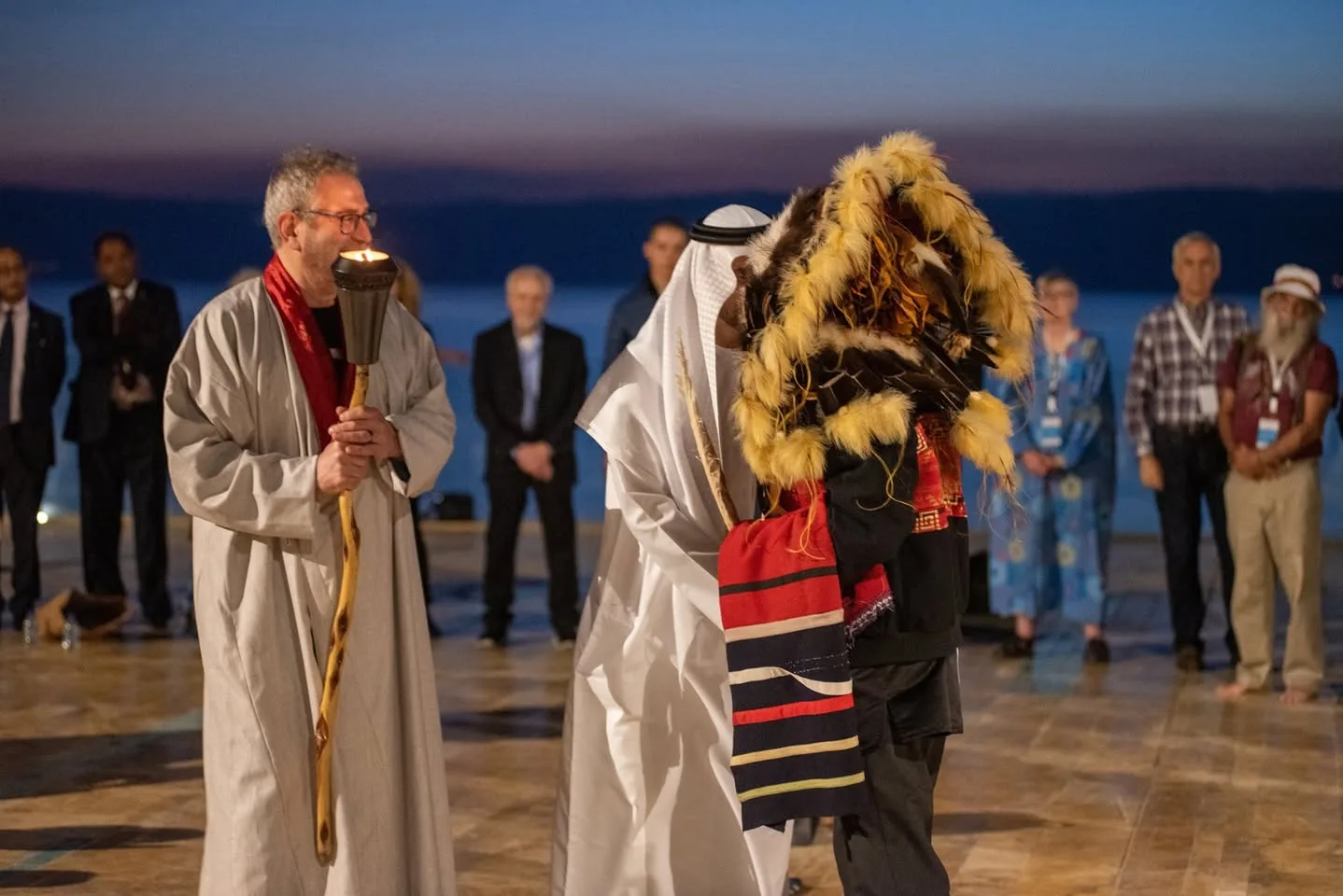 The elder in the feathered headdress embraces the man in white robes as the robed torch bearer smiles and onlookers stand in a circle by the sea at twilight.