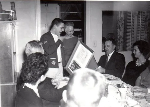 Black-and-white photo of a dinner gathering where a young man in a letter jacket stands beside a woman as a seated man holds up a framed picture.