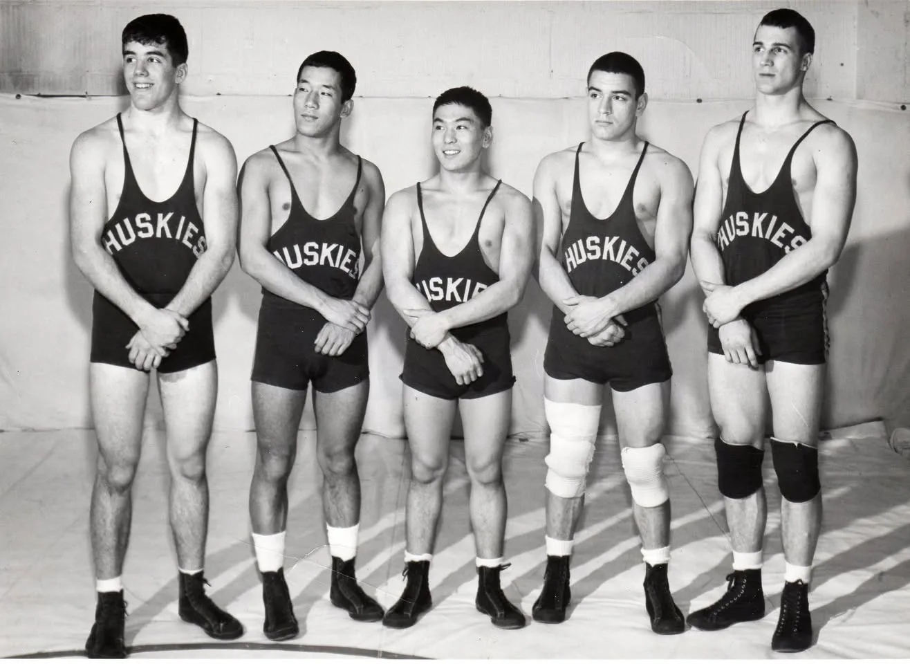 Five young wrestlers in dark Huskies singlets stand in a row on a mat, arms folded in front of them.
