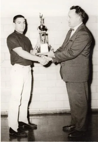 A young man in a dark shirt shakes hands with a man in a suit as they hold a tall trophy together.