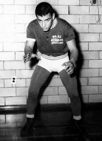 A young wrestler in a Wa-Hi 147 lbs wrestling shirt and tights crouches in a stance before a brick wall.