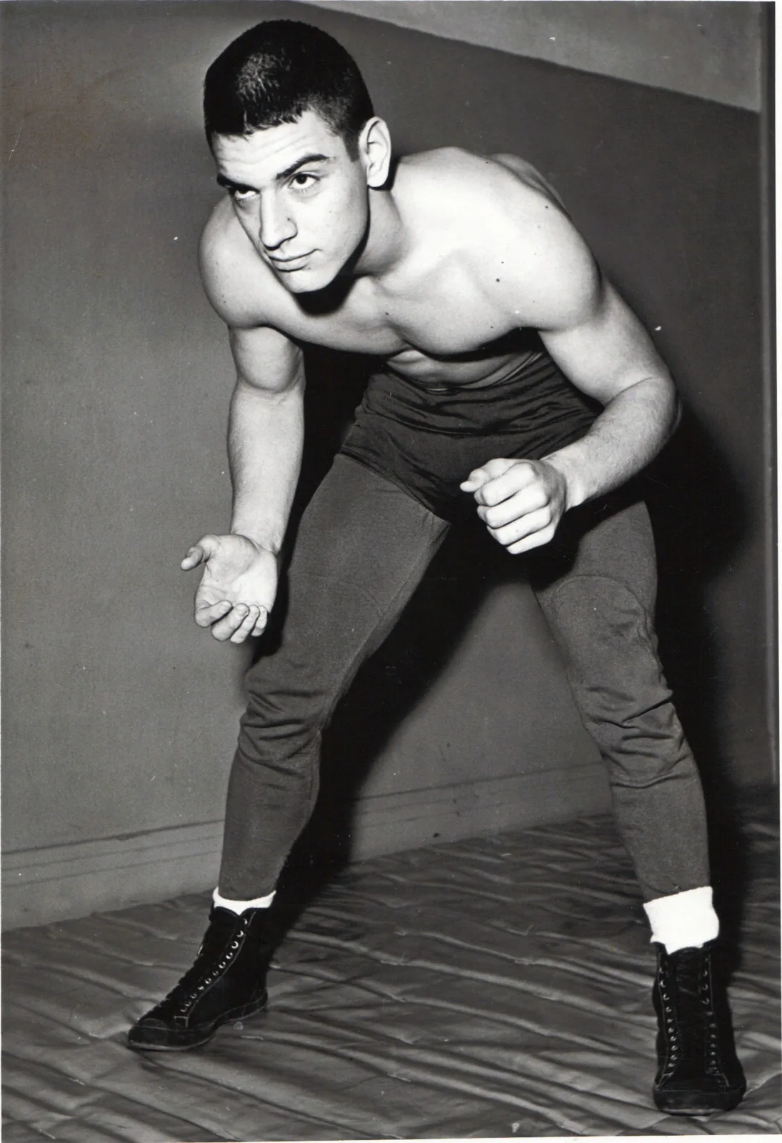 A shirtless young wrestler in dark tights leans forward in a ready stance on a mat.
