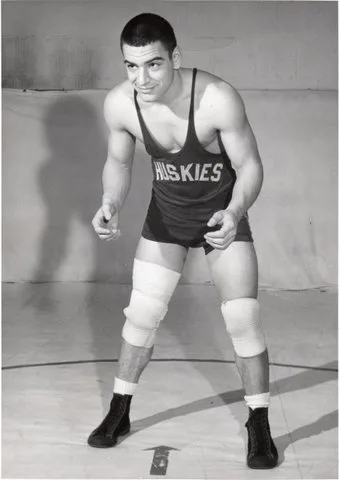 A young wrestler in a Huskies singlet and knee pads crouches in a ready stance on a mat.