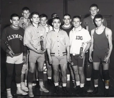 Group of young wrestlers in team jackets and singlets pose holding small trophies.