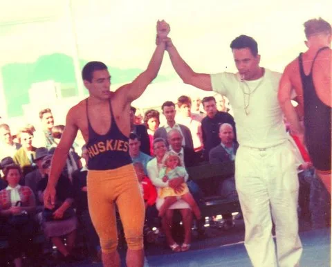Color photo of a referee raising the arm of a wrestler in a Huskies singlet and gold tights in victory before a seated crowd.