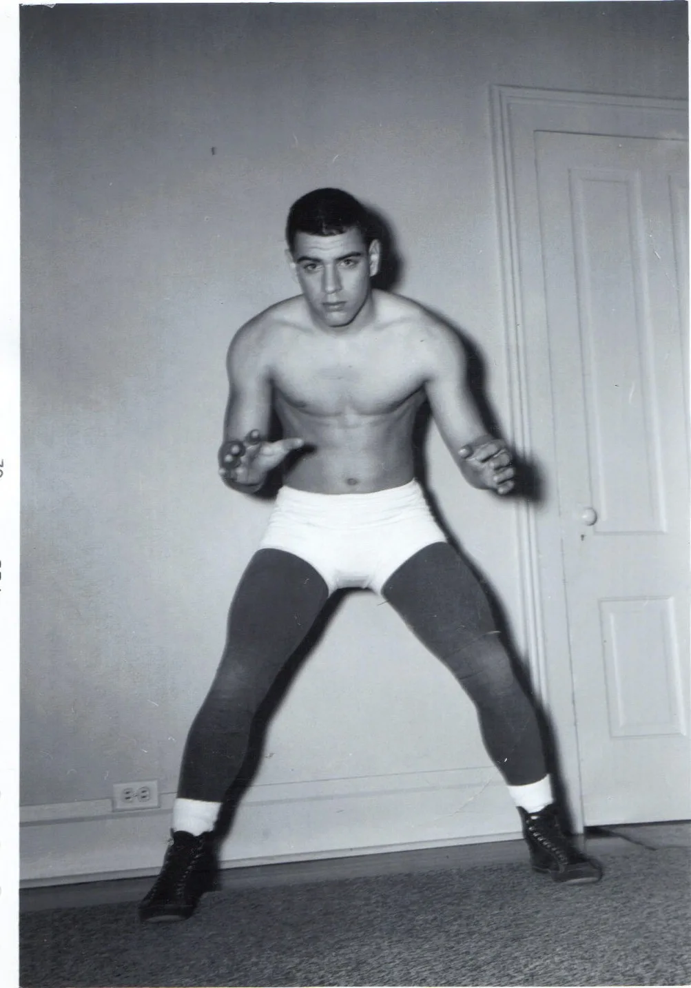 A shirtless young wrestler in white trunks and dark tights crouches in a wide stance in a room beside a door.