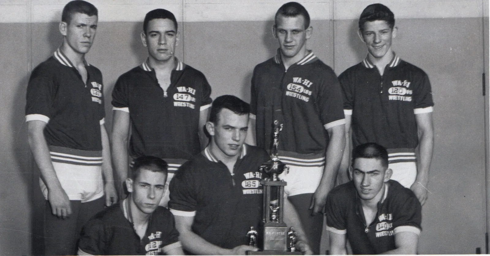 Seven Wa-Hi wrestlers in team shirts pose around a large trophy.