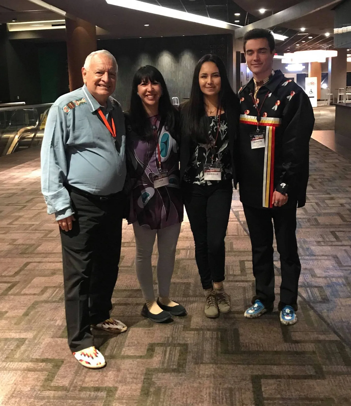 Four people with conference lanyards and name badges pose smiling in a carpeted hall: an elder in a light blue shirt and beaded moccasins, two women, and a young man in a black ribbon shirt.
