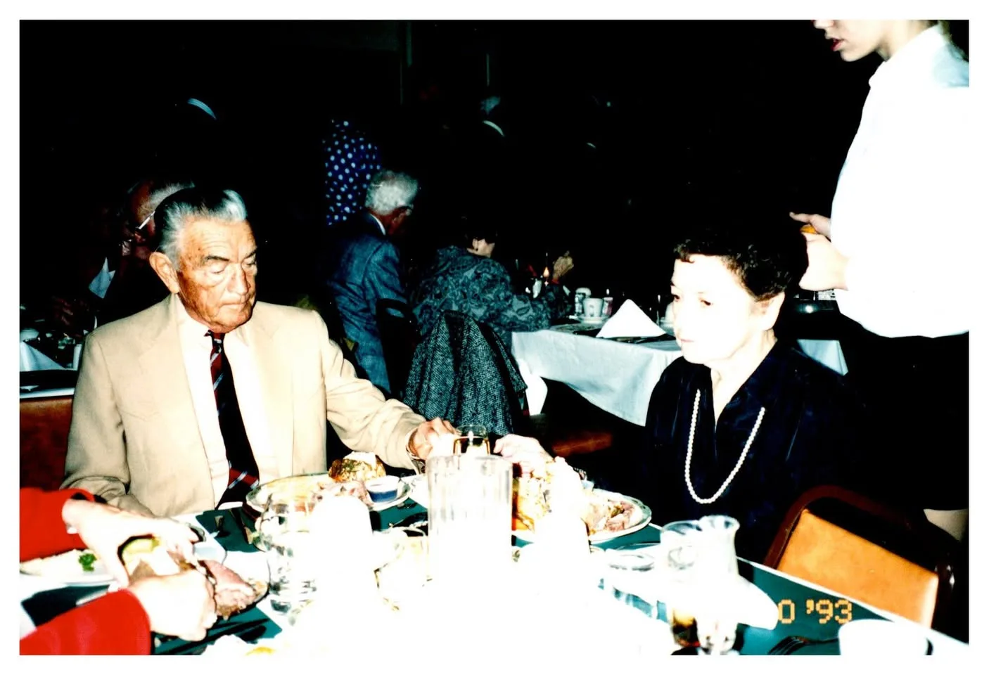 An older man in a tan jacket and a woman in pearls seated at a restaurant table, dated 1993.