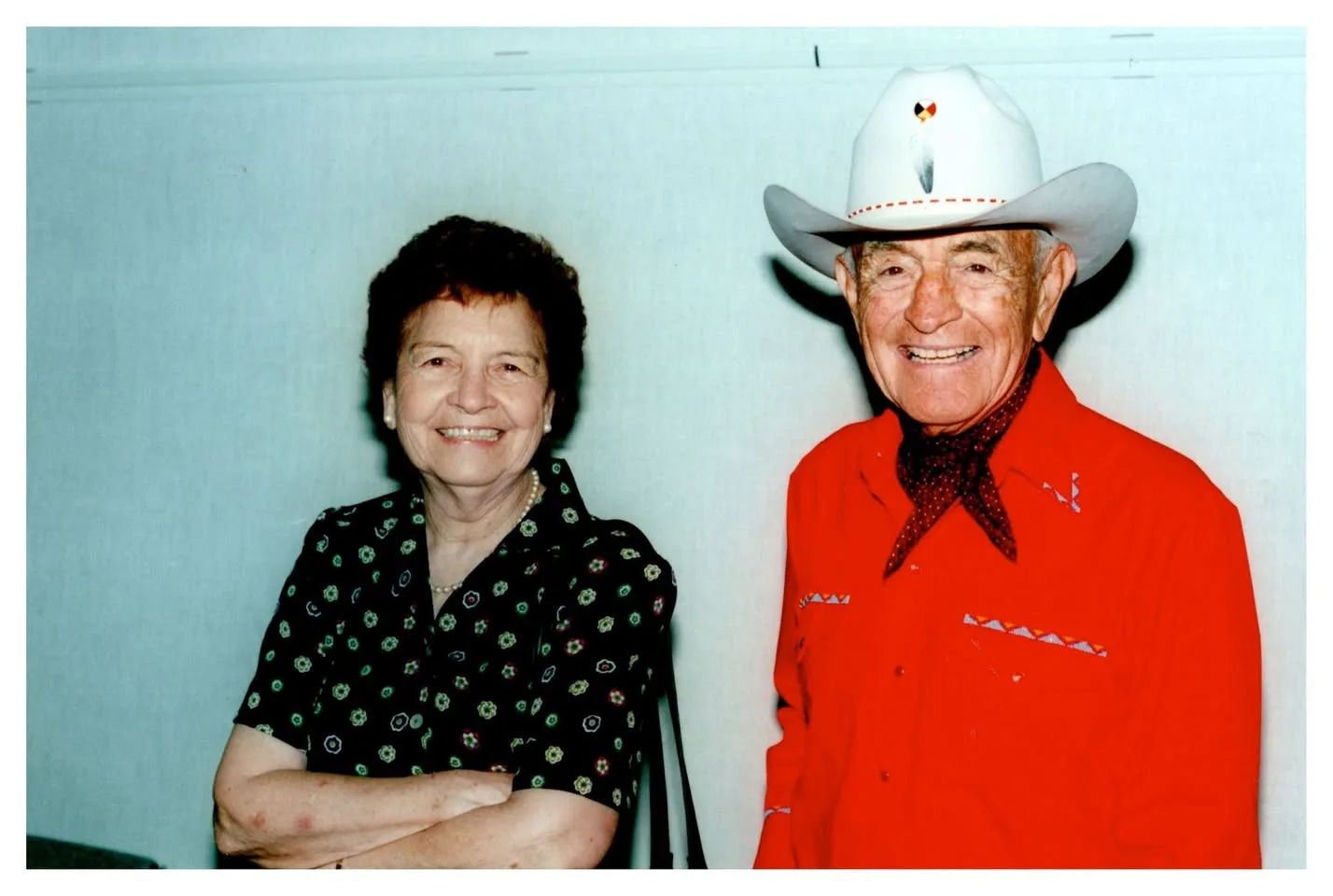 A smiling woman in a floral dress beside a smiling elder in a white cowboy hat and red western shirt.