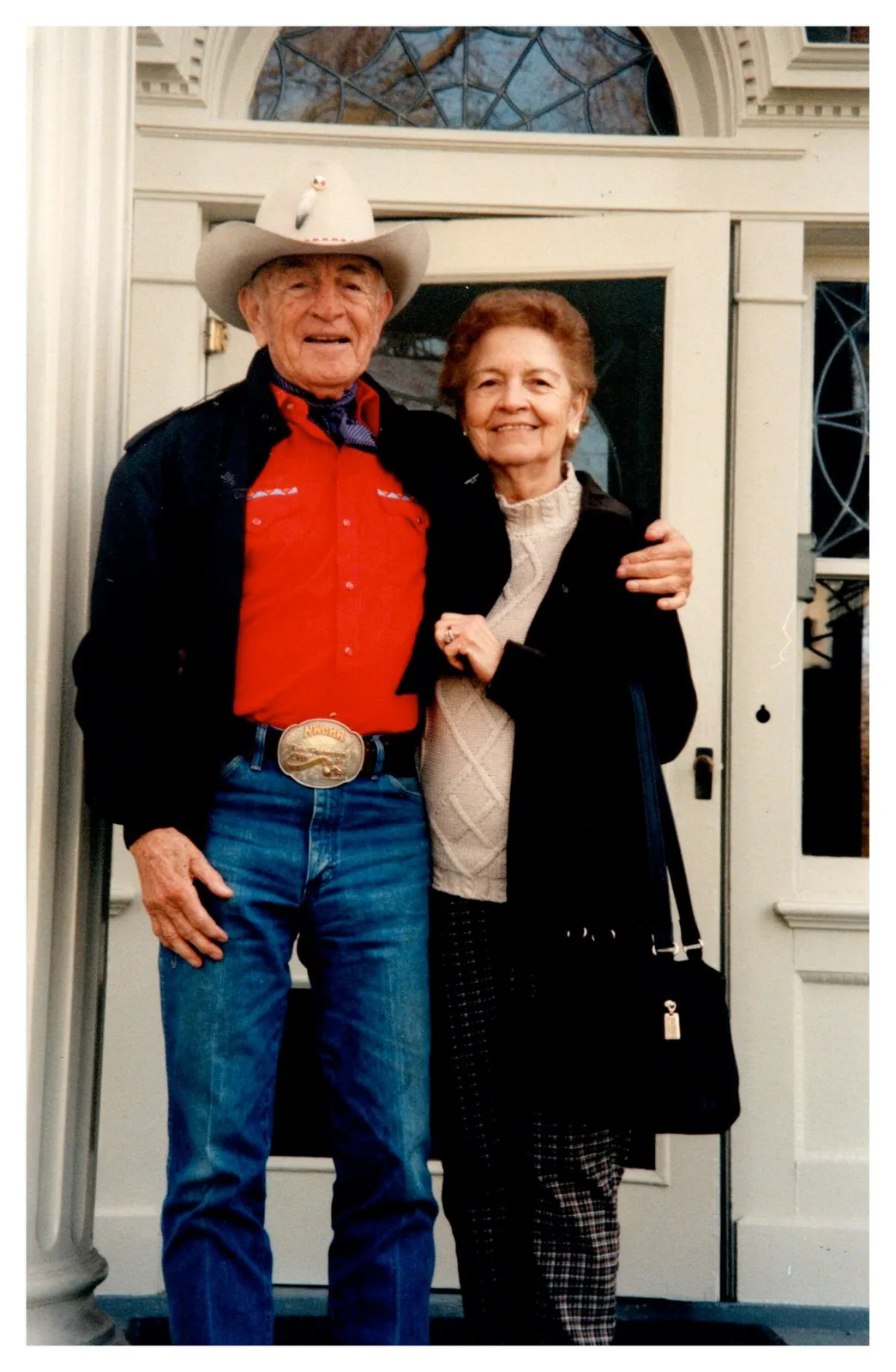 An elder in a cowboy hat and red shirt with his arm around a smiling woman in front of a white doorway.