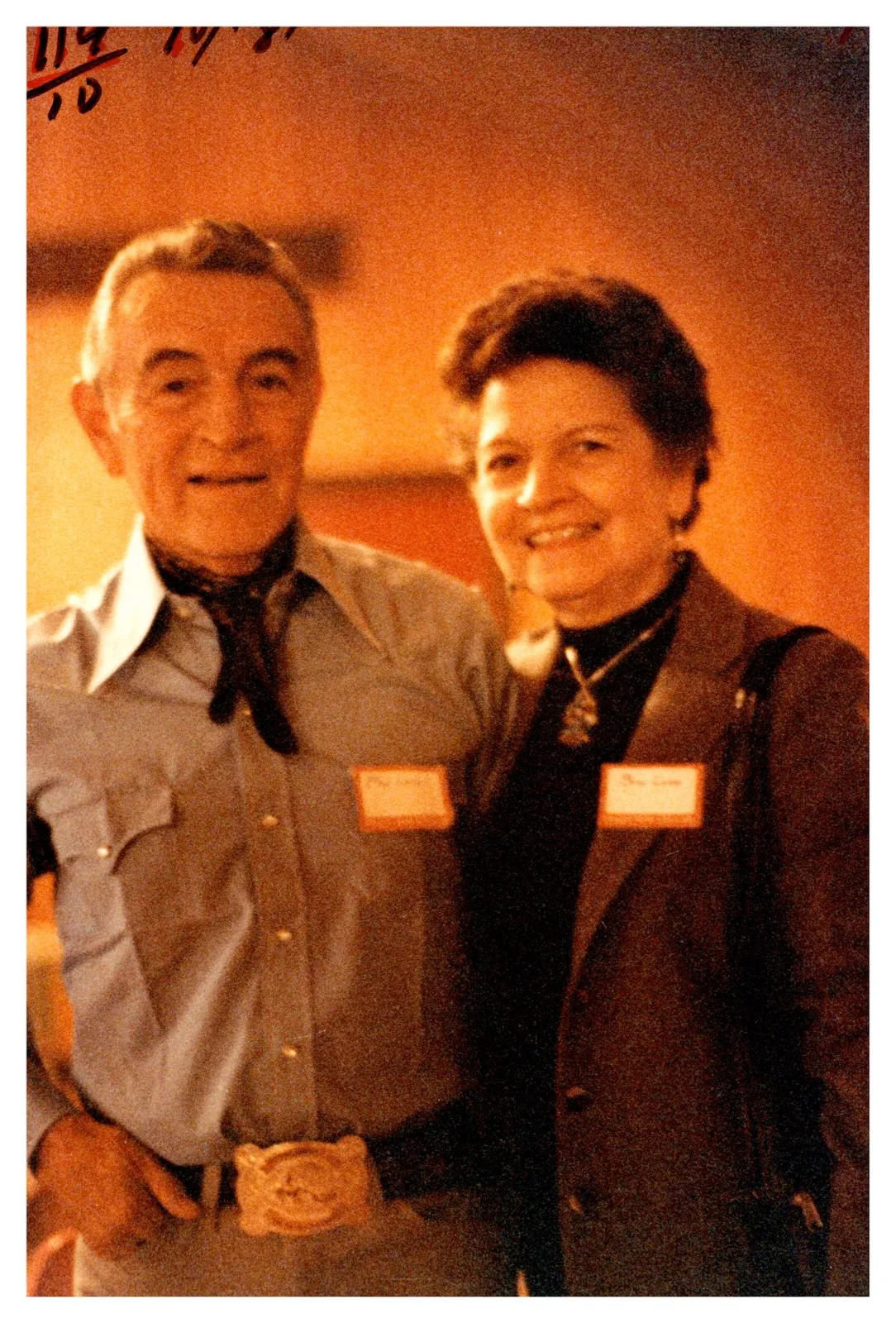 A man in a western shirt and bandana and a woman, both wearing name tags, smiling together.