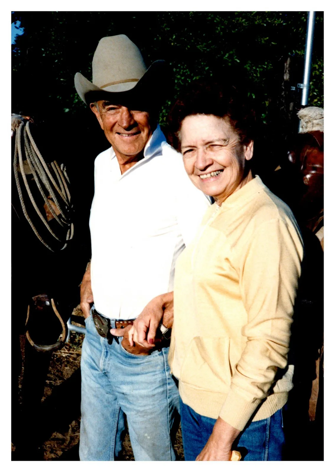 A smiling man in a cowboy hat and white shirt beside a woman in a yellow cardigan, next to a saddled horse with a rope.